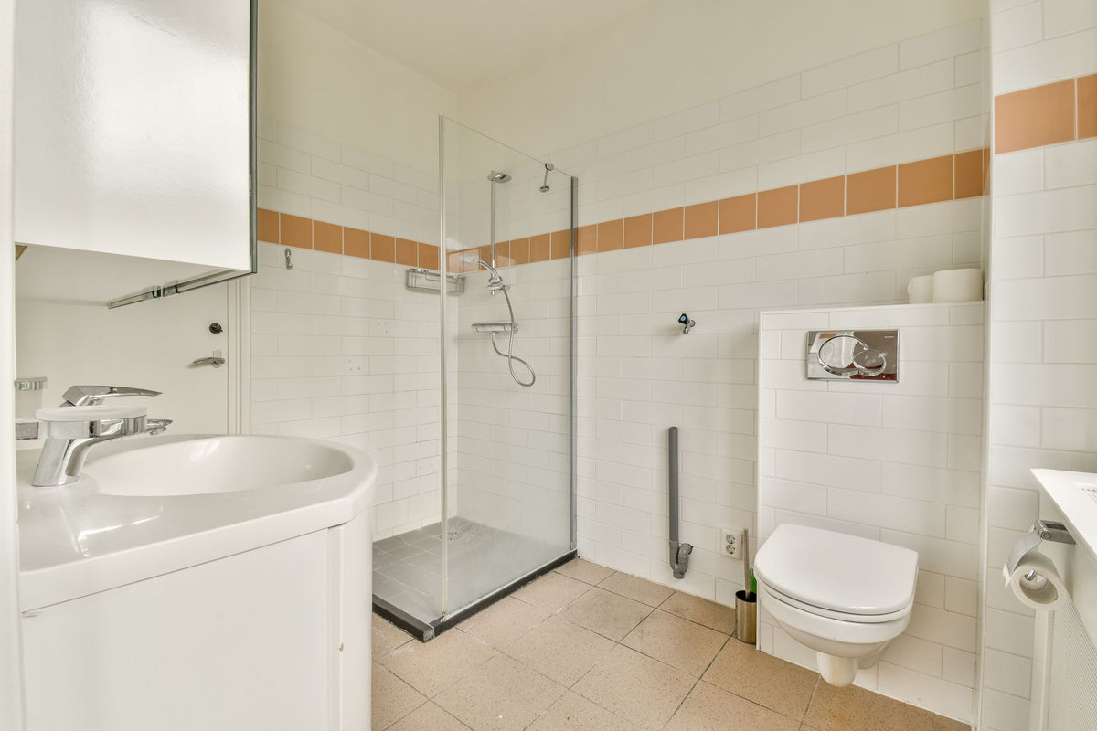Badkamer vóór renovatie met glazen douchewand, witte wandtegels en een oranje tegelrand