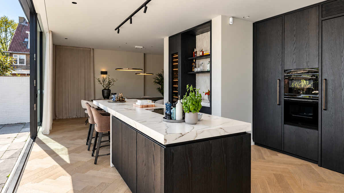 After: luxe maatwerk woonkeuken van Culimaat in een verbouwde jaren ’30 woning in Breda, met donker houten kookeiland, marmerlook werkblad, hoge kastenwand en glazen schuifpui naar de tuin.
