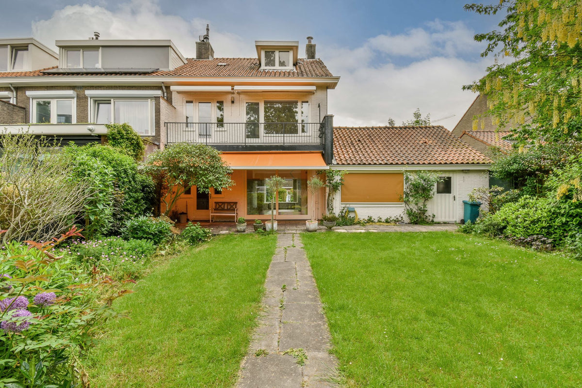 Achtergevel vóór renovatie met een balkon, oranje zonwering en een groene tuin
