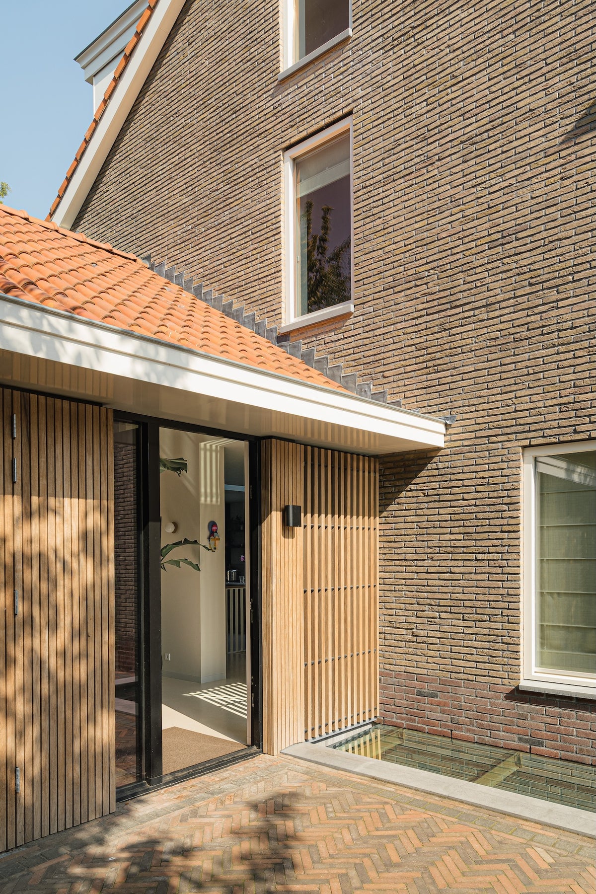 Glazen entree met houten latten naast de bakstenen zijgevel van de woning in de Prinses Irenebuurt