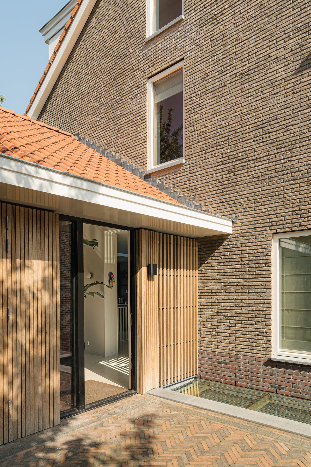 Glazen entree met verticale houten latten naast een bakstenen zijgevel